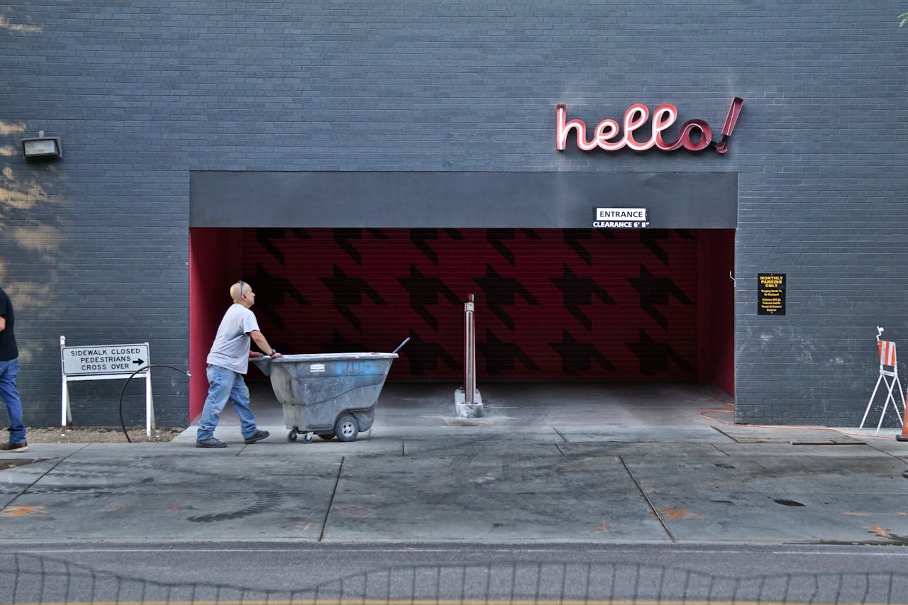 Worker pushes trash cart past a building with 'hello!' sign, highlighting urban life.